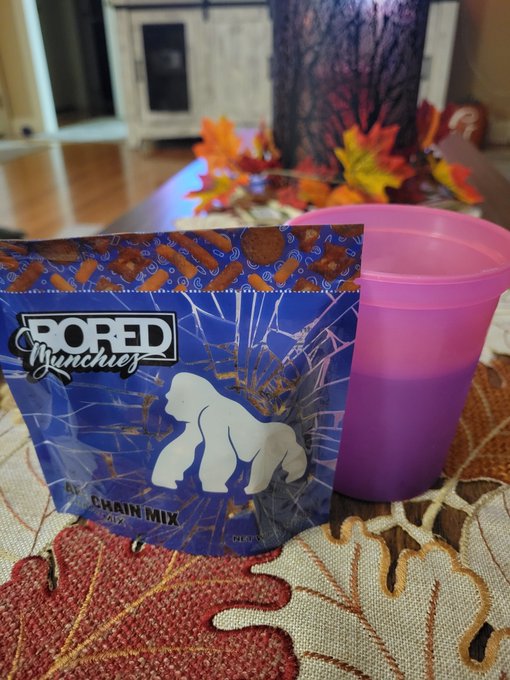 A bag of 'BORED Munchiez' Peanut Train Mix candy sitting next to a pink plastic cup on a table with an autumn leaf patterned tablecloth.