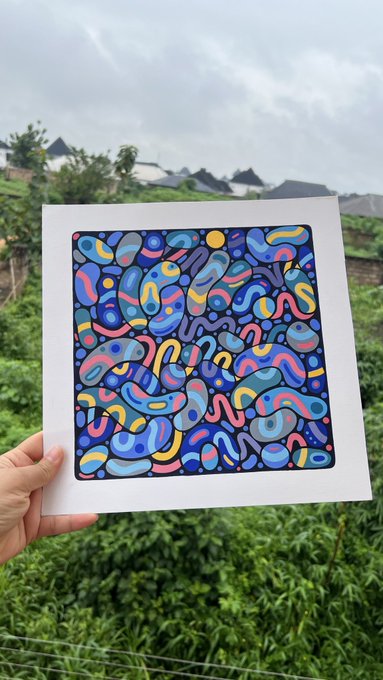 A hand holds up a colorful abstract artwork print featuring swirling blue, pink, yellow, and purple organic shapes against a backdrop of green foliage and an overcast sky.