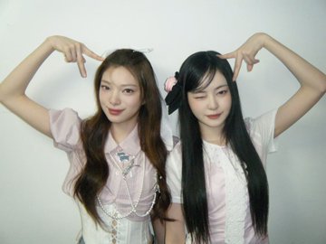 Two young women posing together against a white wall, both forming heart shapes with their arms above their heads; one has long brown hair in a pink outfit and the other has long black hair with a pink bow in a white and pink dress.