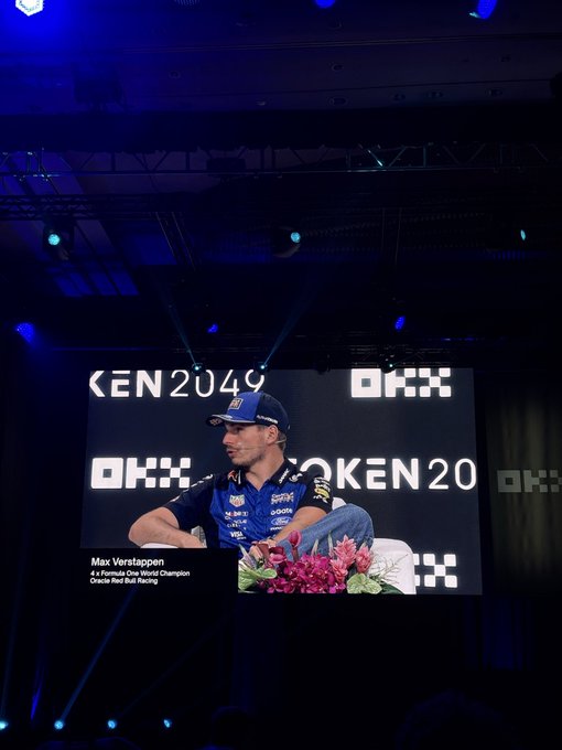 A large stage screen displaying an image of Max Verstappen, identified as a 4x Formula One World Champion, with 'OKX' and 'TOKEN2049' branding.