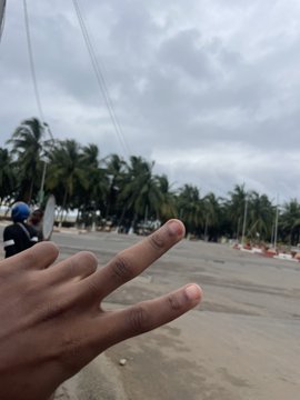 A hand makes a peace sign in the foreground of an empty street lined with palm trees under a cloudy sky.