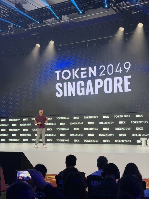 A speaker presents on stage at TOKEN2049 Singapore, with the event's branding and "OKX" sponsor logos prominently displayed on a massive backdrop, contextualizing Haseeb's talk on AI and privacy as part of a major crypto conference.