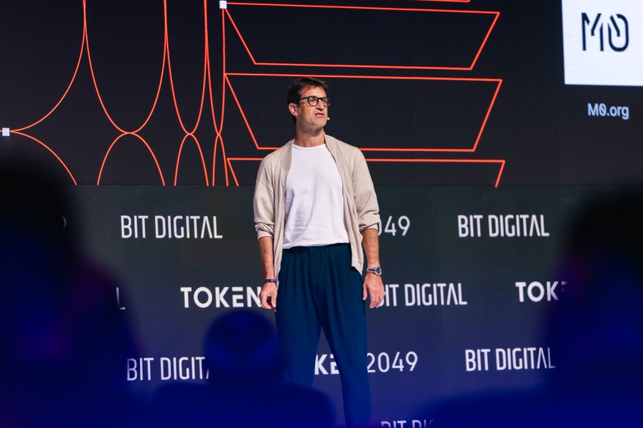 A man wearing glasses, a white shirt, and blue pants stands on a stage with 'BIT DIGITAL' and 'TOKEN 2049' branding visible on the backdrop.