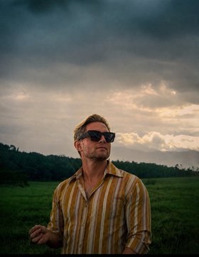 A man in a striped yellow shirt and sunglasses gazes upward against a dramatic cloudy sky, projecting quiet confidence and visionary focus. The image visually reinforces the post's message of mysterious ambition—"You don't understand what I am building"—suggesting the ticker **$TBB** represents something others haven't yet grasped or appreciated.