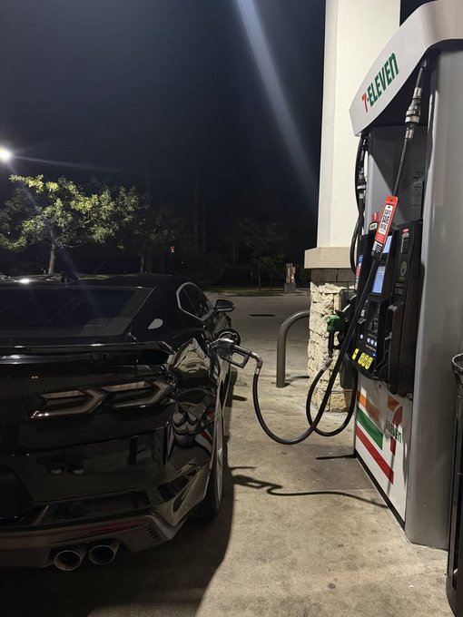 A nighttime photo of a black Lamborghini Huracán being refueled at a 7-Eleven gas station, showing the car's rear and the illuminated fuel pump.