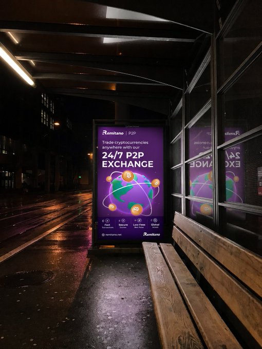 A glowing Remitano P2P advertisement in a nighttime bus shelter advertises "24/7 P2P EXCHANGE" for trading cryptocurrencies, perfectly illustrating the caption's message that the platform is available anytime, rain or shine, day or night. The vibrant purple billboard, visible against the wet, dark street, reinforces the always-on, anywhere accessibility of the crypto trading service.