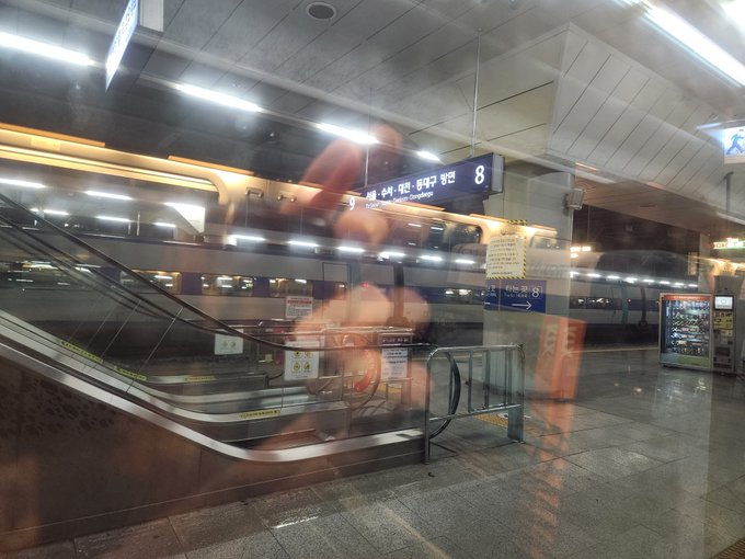 A slightly blurred photo taken through glass at a Korean subway station showing an escalator, with signage indicating directions for 'Gangnam' and 'Jangchungdanneung' at exit 8.