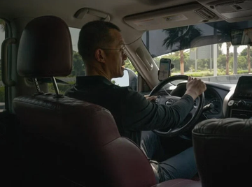 A man wearing glasses and a dark long-sleeved shirt sitting in the driver's seat of a vehicle, with his other hand reaching toward the dashboard area.