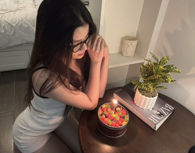 A woman with glasses sits at a small table with a lit candle surrounded by colorful stones in a bowl, next to a potted plant and a 'BAZAAR' magazine.