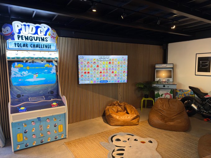 A Pudgy Penguins "Polar Challenge" arcade game and a "Wall of Penguins" digital display sit in a lounge area with bean bag chairs, embodying the brand's new community-focused initiative called "The Huddle."