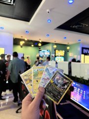 A person's hand holding up four Pokémon trading cards inside a brightly lit convention hall or store, with other attendees and booths visible in the blurred background.