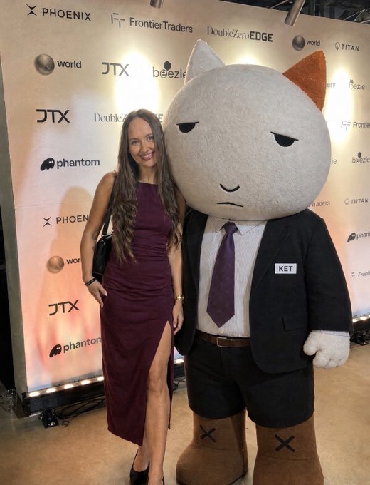 A woman in a dark red/maroon dress poses next to a large mascot character wearing a suit and tie with a 'KET' name tag, at an event with sponsor logos including Phoenix, FrontierTraders, DoubleZero EDGE, JTX, and Phantom.