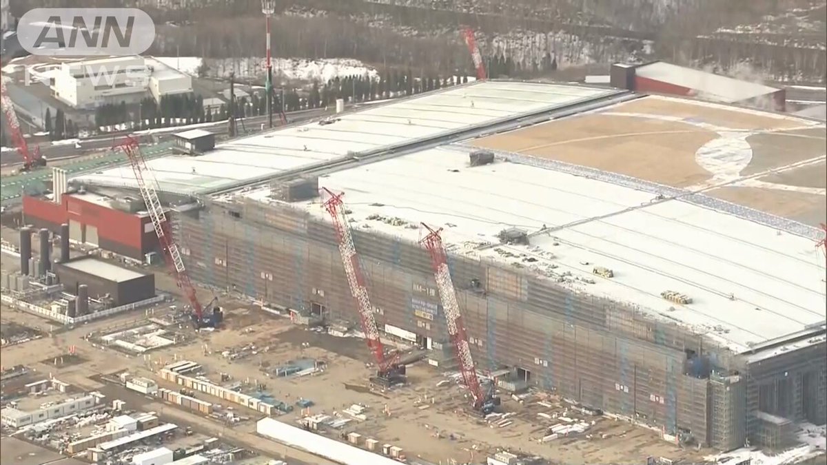 The image shows a large building under construction with several cranes working on different parts of the structure. The crane operators are wearing hard hats for safety while they work. There are also people present at various locations around the site, likely overseeing the progress or assisting in the construction process. The scene captures the scale and complexity of the ongoing project as it unfolds over time.
