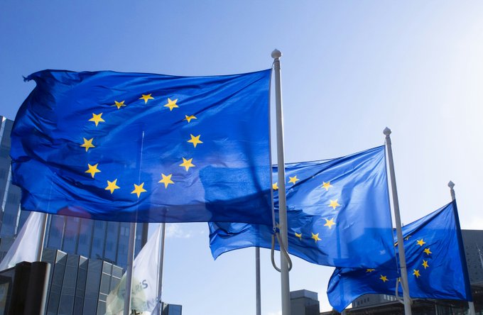 Multiple European Union flags wave against a clear blue sky outside a modern building, symbolizing the EU regulators referenced in the accompanying post about warning customers against scammers impersonating officials to steal crypto assets.