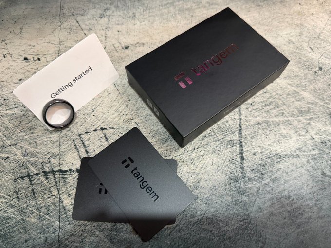 Tangem card wallet unboxed: the package includes the branded Tangem box, "Getting started" guide, two black Tangem smart cards, and a ring, all arranged on a scratched concrete surface.