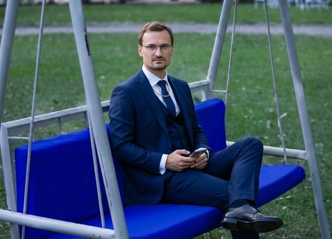 A man in a navy suit and glasses sits on a blue swing seat outdoors, holding a phone.