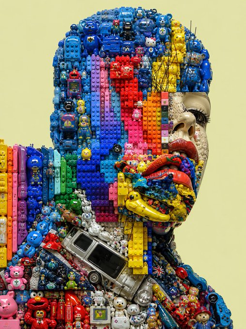 A mixed-media portrait of a face composed of colorful plastic toys, building blocks, figurines, and a small toy bus against a yellow background.