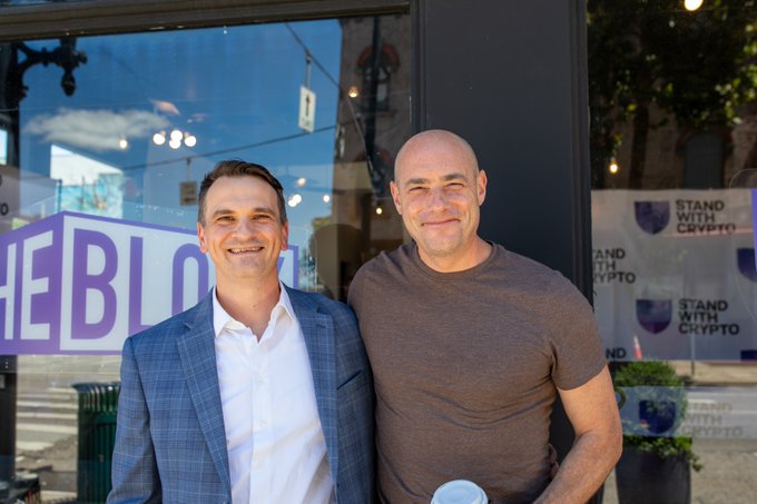 Two men pose together at an outdoor event, with one wearing a blue blazer and the other in a gray t-shirt, standing in front of glass displaying "Stand With Crypto" signage and partial text reading "HE BLOCK." The image captures the SWC Community Roundtable event where Congressman Rep. Greg Landsman joined attendees for "On The Block," a series of discussions about how digital assets can benefit local communities through reduced costs and increased access to capital.