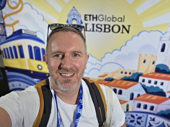 A selfie of a smiling man with sunglasses on his head and a backpack, standing in front of a colorful illustrated backdrop for the 'ETHGlobal LISBON' event.