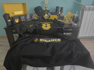 A celebratory display of Binance and BNB Chain merchandise including apparel, shoes, and accessories arranged on a bed, commemorating the platforms' 9th anniversary with branded items prominently featuring the "BINANCE" logo and the signature yellow and black color scheme.