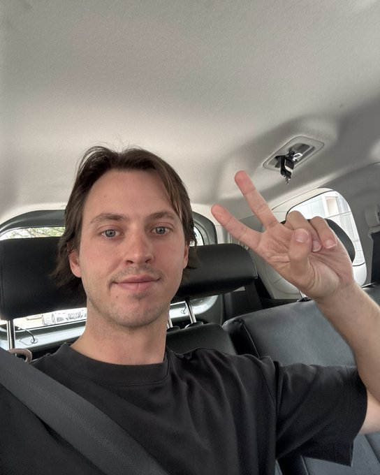 A selfie of a young man with medium-length brown hair sitting in a car, wearing a black t-shirt and seatbelt, making a peace sign with his fingers.