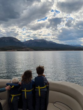 A scenic photograph of three children wearing yellow life jackets sitting on a boat, looking out at a lake with mountains and a dramatic cloudy sky in the background.