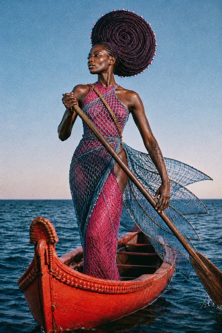 An editorial fashion photograph of a woman standing in a red wooden boat on the ocean, wearing a pink mesh gown with blue flowing fabric and a large coiled purple headpiece.