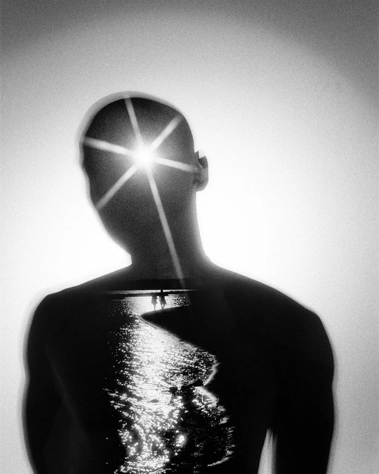 A surreal black-and-white double exposure artwork showing a man's silhouette merged with a sunset over water.