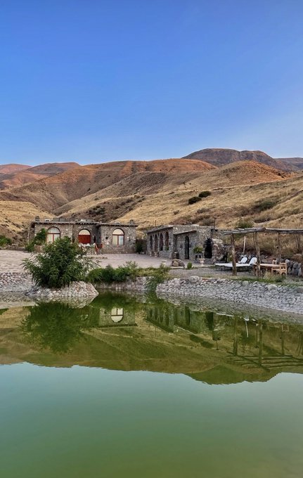 A scenic landscape showing a stone building complex with arched doorways reflected in a calm green pond, set against a backdrop of arid, rolling brown mountains under a clear blue sky.