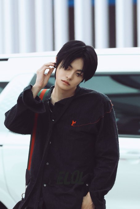 A young man with dark hair, likely a K-pop idol, stands outdoors near a white car wearing a black corduroy jacket with red accents, adjusting his hair; the image is watermarked with 'ELOL'.