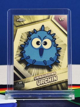 A "VF Unwavering Urchin True Super" VeeFriends trading card, featuring a blue spiky cartoon urchin with big eyes on a gold background, displayed in a clear protective case. The card showcases the Topps Chrome logo, "VF" branding, and the "Unwavering Urchin" character name—fittingly shared in the post about photographing this Super Scene Swap proof card.