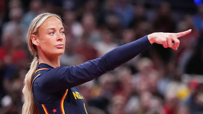 A blonde female athlete wearing a dark long-sleeved jersey with red stars pointing outward, appearing to be directing teammates during a sporting event.