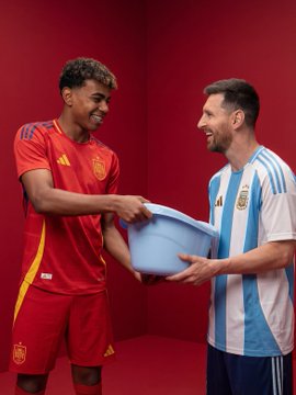 Two soccer players wearing Spain and Argentina national team kits smiling and exchanging a light blue jersey in a red studio setting.