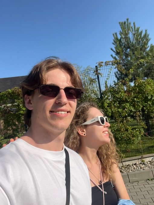 A young man in sunglasses and a white t-shirt and a young woman in white Celine sunglasses smile together in a sunny outdoor setting, with the man visibly wearing the "$ANSEM" energy as he returns from a refreshing weekend log-off.