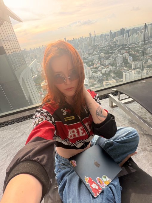 A red-haired woman wearing sunglasses, a checkered racing-style jacket, and baggy jeans poses on a rooftop with a city skyline at sunset, holding an Apple laptop covered in stickers.
