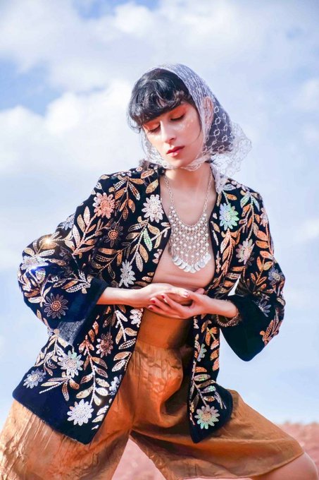 A fashion photograph of a woman with bangs wearing a sheer veil and an ornate floral-embroidered dark jacket over a bronze skirt, posing against a cloudy sky and desert landscape.