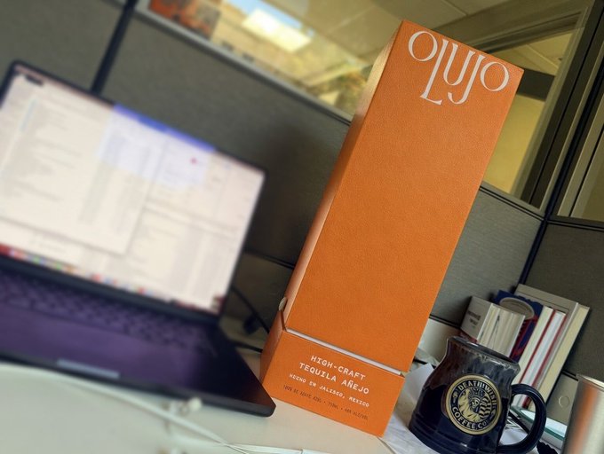 An orange box of 'Ojo Ljo' High-Craft Tequila Añejo from Valle de Jalisco, Mexico, sitting on a desk next to a laptop and a black coffee mug.