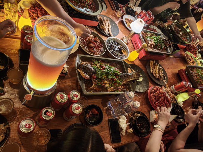 A dimly lit dining table is filled with various Asian dishes including a whole fish in sauce, crayfish, and skewers, alongside cans of Tsingtao beer and a glowing beverage dispenser.