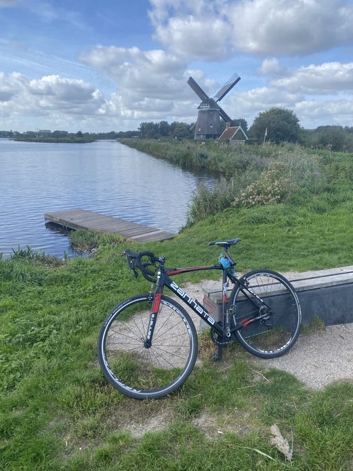 A black, red, and silver Giant road bicycle leans against a low wall on a grassy path beside a calm canal, with a traditional windmill and a small wooden dock visible in the background under a cloudy sky.