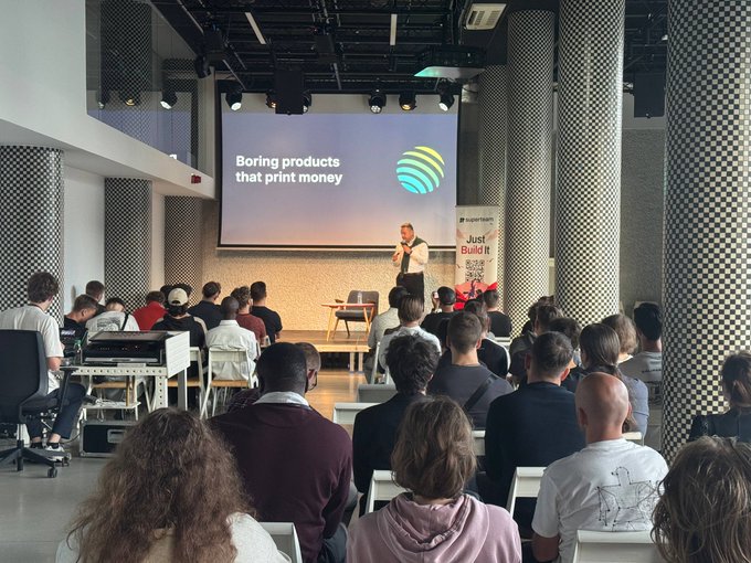 A conference presentation in a modern venue with checkered columns, where a speaker stands before a screen displaying 'Boring products that print money' alongside an audience.