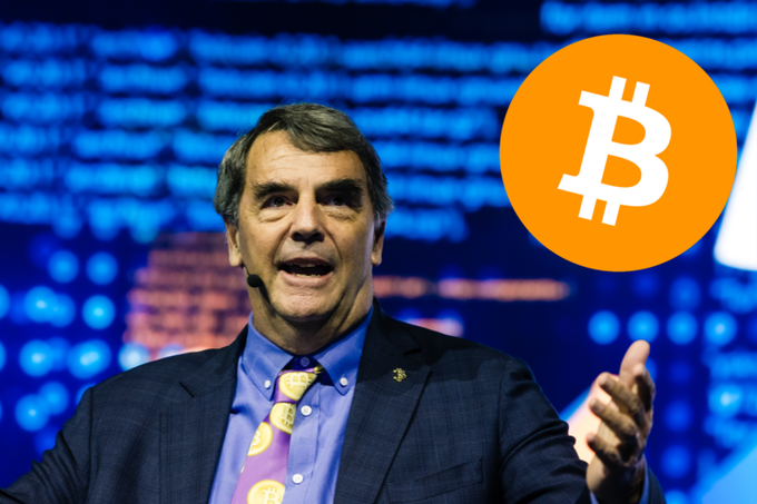 A man in a blue suit and Bitcoin-patterned tie speaks on a stage with a blue digital background, accompanied by a large orange Bitcoin logo graphic.