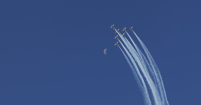 A formation of approximately six jets flies across a clear blue sky, leaving white smoke trails, with a crescent moon visible in the background.