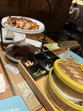Table at a dim-sum restaurant featuring a tiered serving stand with sliced roasted meats, a bamboo steamer, rice, dipping sauces, and a black condiment tray.