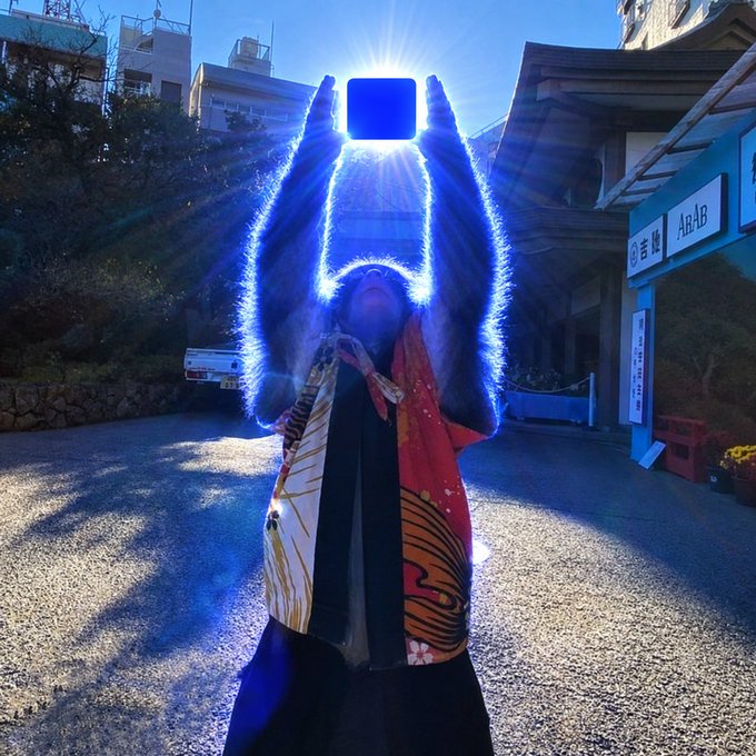 A silhouetted figure in a kimono raises a glowing cube toward the sun, creating a dramatic blue halo of light against a Japanese temple setting.