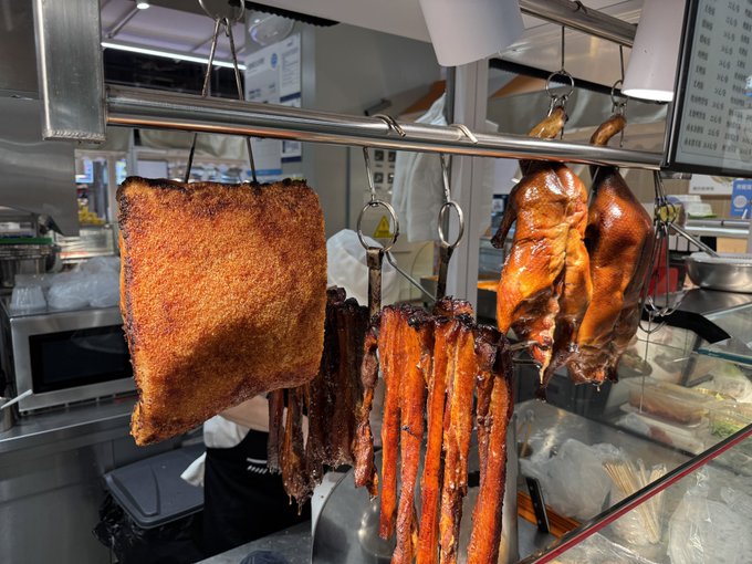 A display of Chinese roasted meats hanging from metal hooks, featuring crispy roast pork, char siu, and a whole roasted duck or goose, with a price list visible in the background.