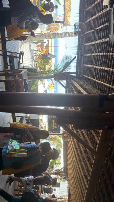 A portrait-oriented photo of a casual restaurant or beach club with a thatched roof, where people are seated at tables and watching a projection screen displaying what appears to be a UEFA Euro match (Hungary vs. Portugal).