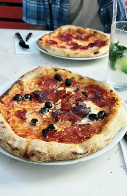 Two personal-sized pizzas served on white plates at a restaurant table, with the foreground pizza topped with pepperoni, olives, and red onions, accompanied by a glass of water with mint.