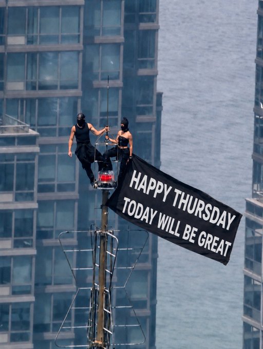 Two people in black outfits and masks perched on a tall pole/antenna high above a city with skyscrapers, holding a black banner that reads 'HAPPY THURSDAY TODAY WILL BE GREAT'.