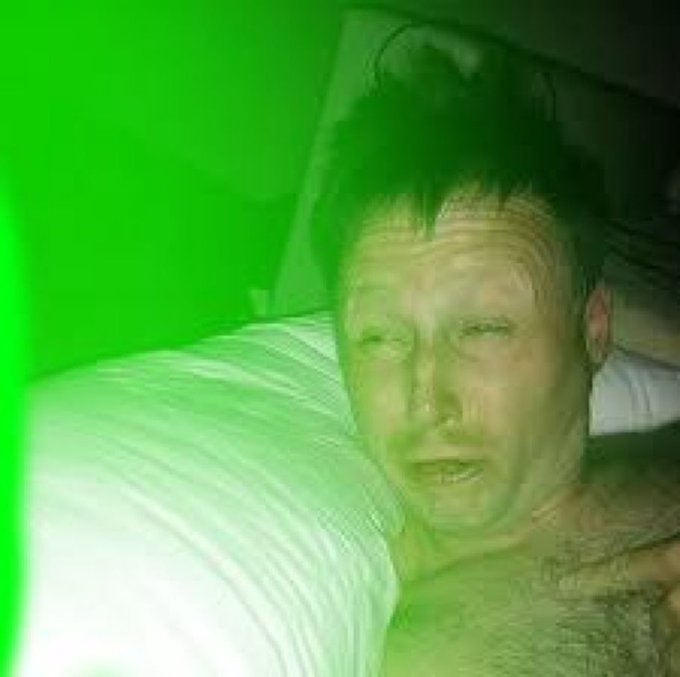 A grainy photo with a strong green tint shows a shirtless man with short dark hair lying against white pillows, appearing weary with his mouth slightly open.