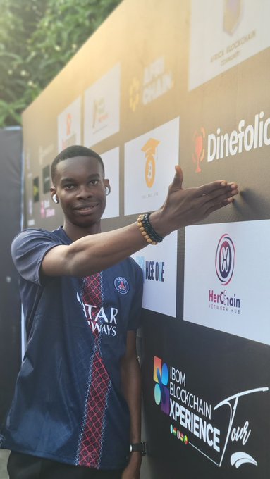 A young man wearing a Paris Saint-Germain 'Qatar Airways' jersey poses with his arm extended in front of a sponsor backdrop featuring logos including 'DineFolic', 'HerChain', 'NEOVE', and 'BOM BLOCKCHAIN XPERIENCE Tour'.